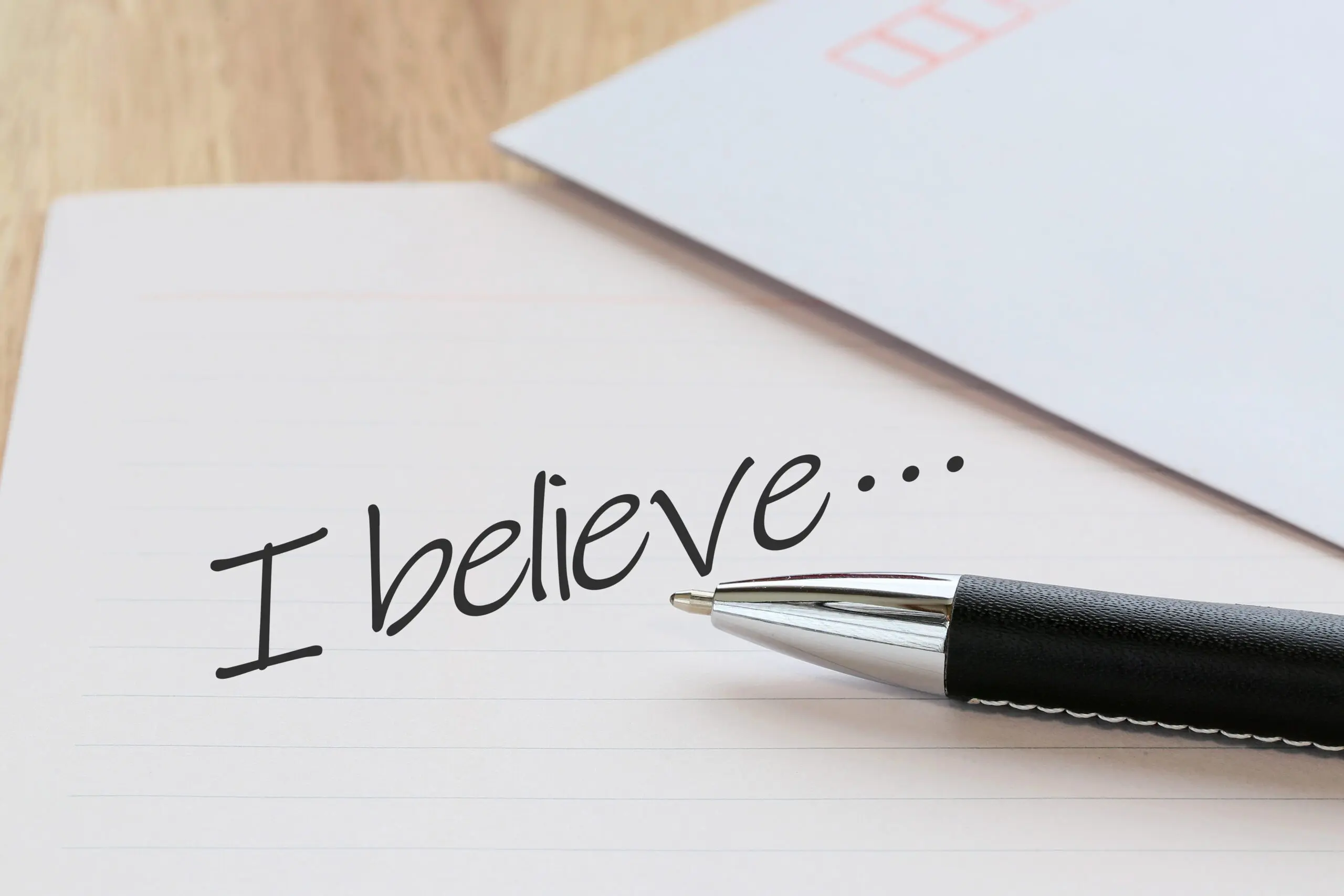 罫線入りの紙の上に銀と黒のペンが置かれ、黒インクで「私は信じています...」と書かれています。これは、英語能力がキャリアアップと転職の成功への新しい道を切り開くことができる旅の始まりを象徴しています。.
