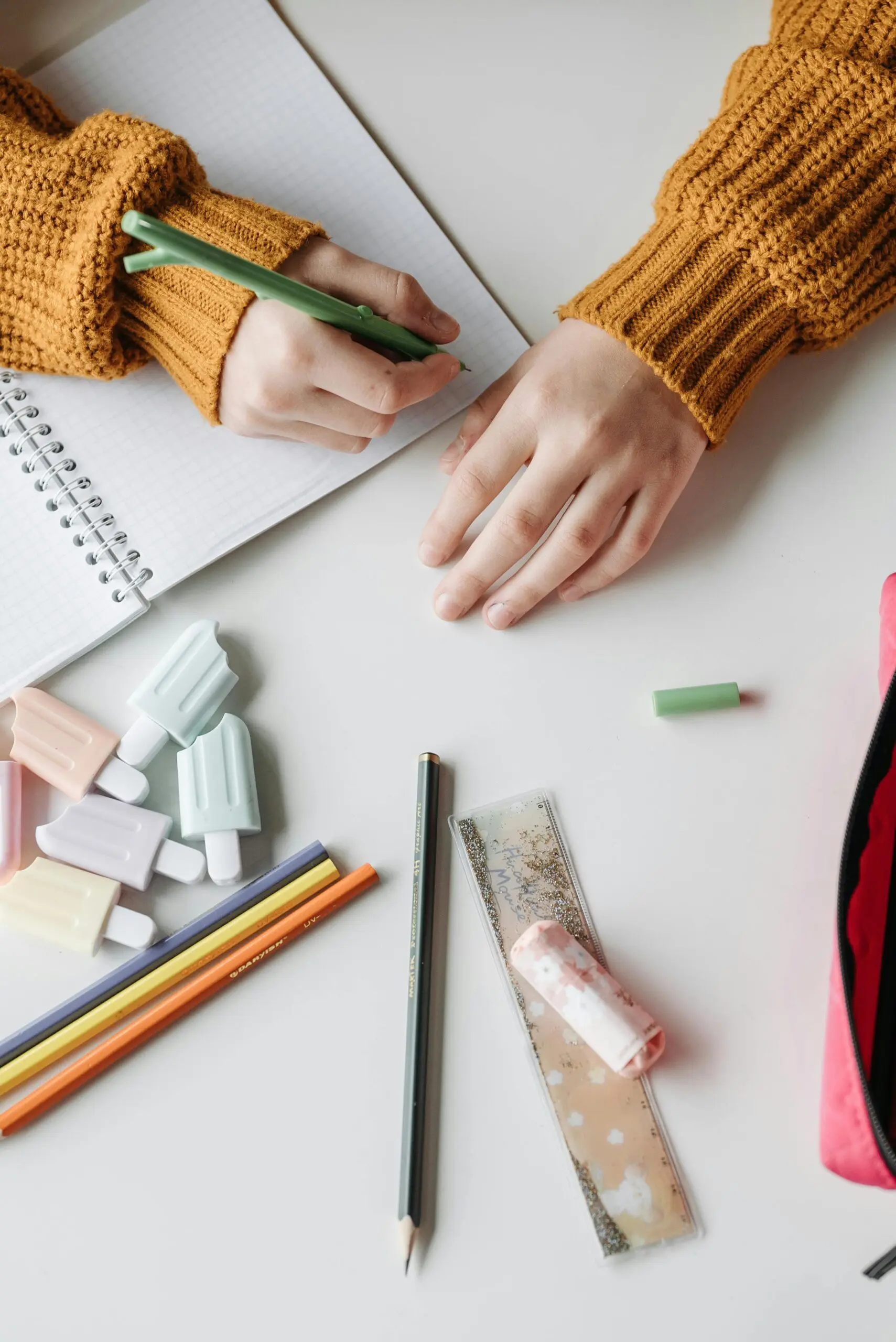 黄色いセーターを着た人が、机の上の鉛筆、カラフルな消しゴム、定規、筆箱に囲まれてノートに書き込み、近々開催される IT 勉強会に向けてスキル向上を図っています。.