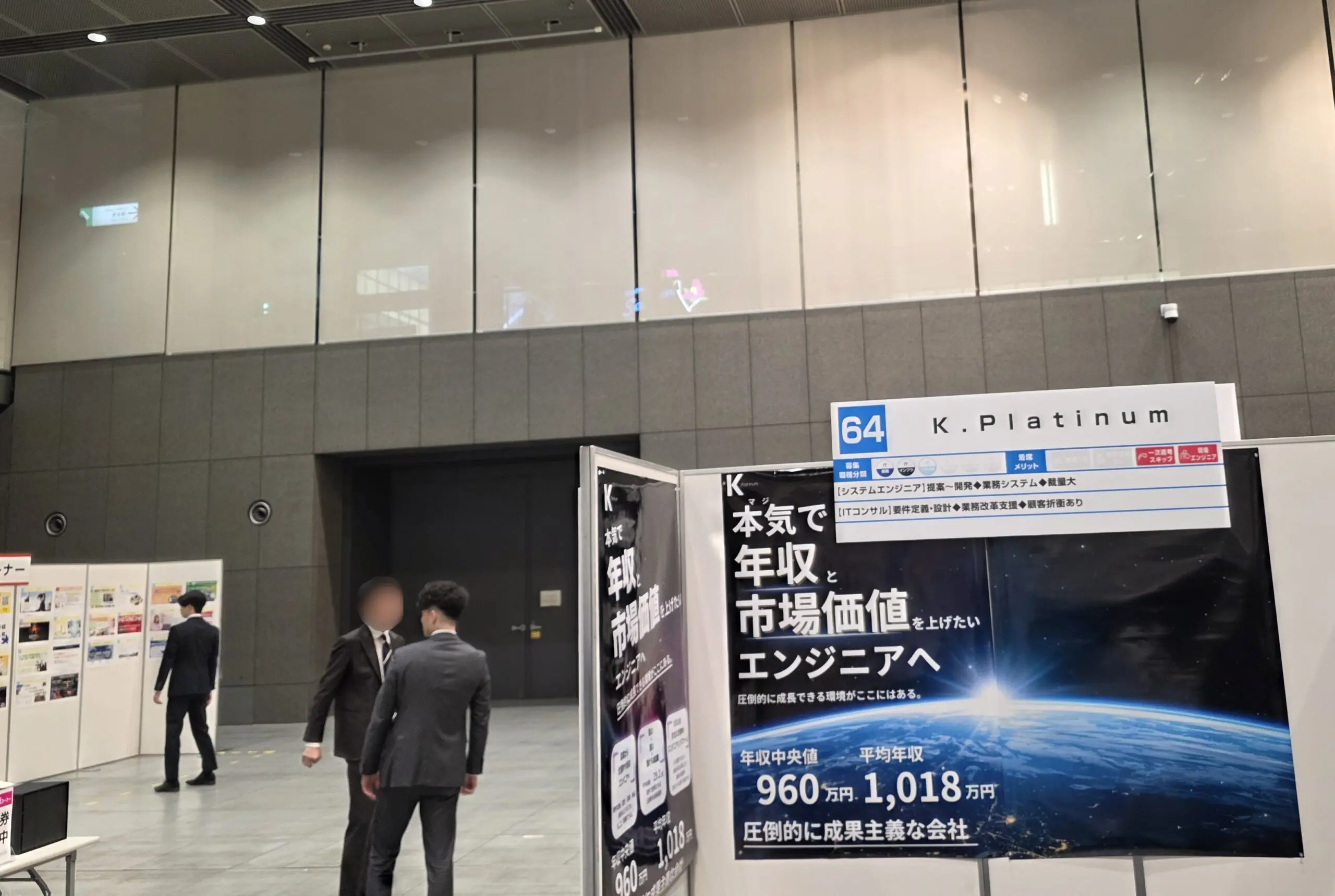 近代的な屋内会場に、日本語のポスターが貼られた「K.Platinum」と書かれたリクルートブース。スーツ姿の男性2人がブースの近くで話している。.