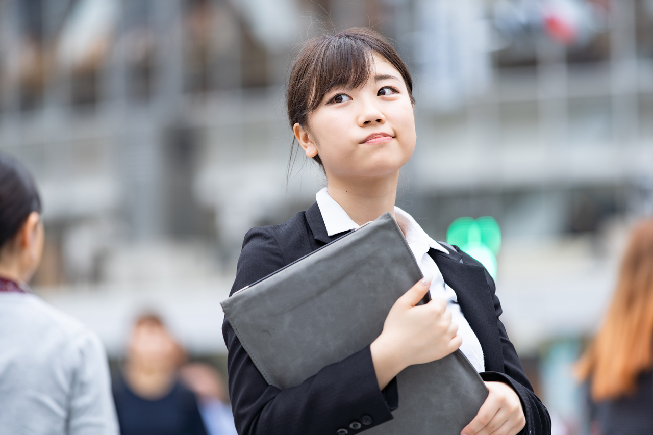 ビジネススーツを着た人物が、フォルダーを手に、背景がぼやけた都市部で面接の準備をしている。.