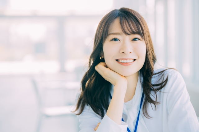 明るくぼやけた室内で、長い茶色の髪の女性が、顎を手に乗せて思慮深く微笑み、年収アップにつながる刺激的な転職を考えています。.