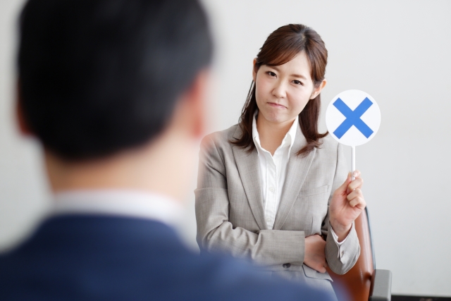 スーツを着た女性が、オンラインプログラミングスクールのミーティングの場で、男性を見つめながら「不採用」を示す青い「X」の看板を掲げている。.