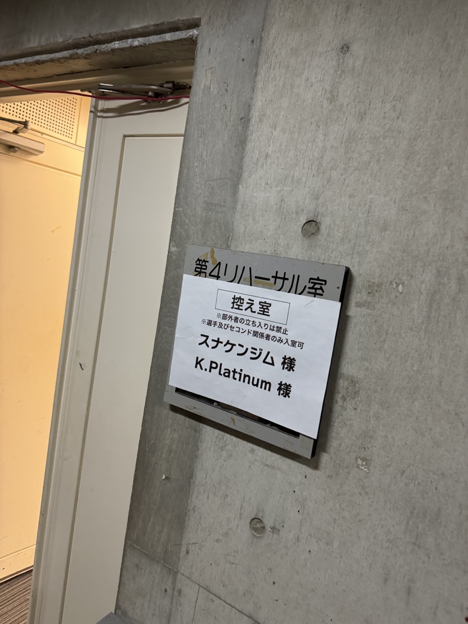 コンクリートの壁にはリハーサル室を示す「第4リハーサル室 控え室スケジュール様 K.Platinum様」と日本語で書かれた看板。左側に部分的に見えるドアは、パフォーマーがレキオバトル名護の興奮の中で参戦する準備をしている間、来たる熱戦を暗示しています。.