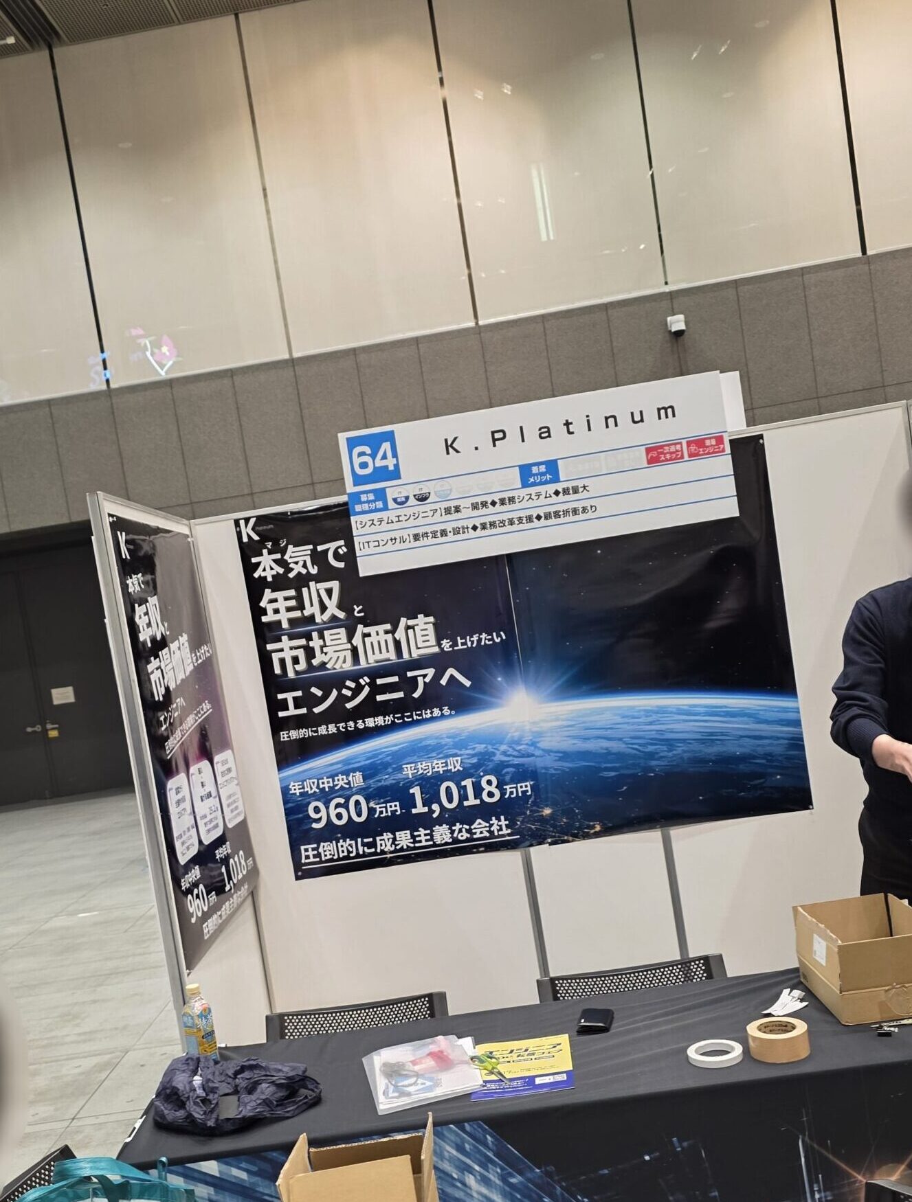 K.プラチナと書かれた展示会ブース。テーブルの上にポスター、箱、テープ、バッグが置かれ、立っている人と座っている人の2人がいる。.
