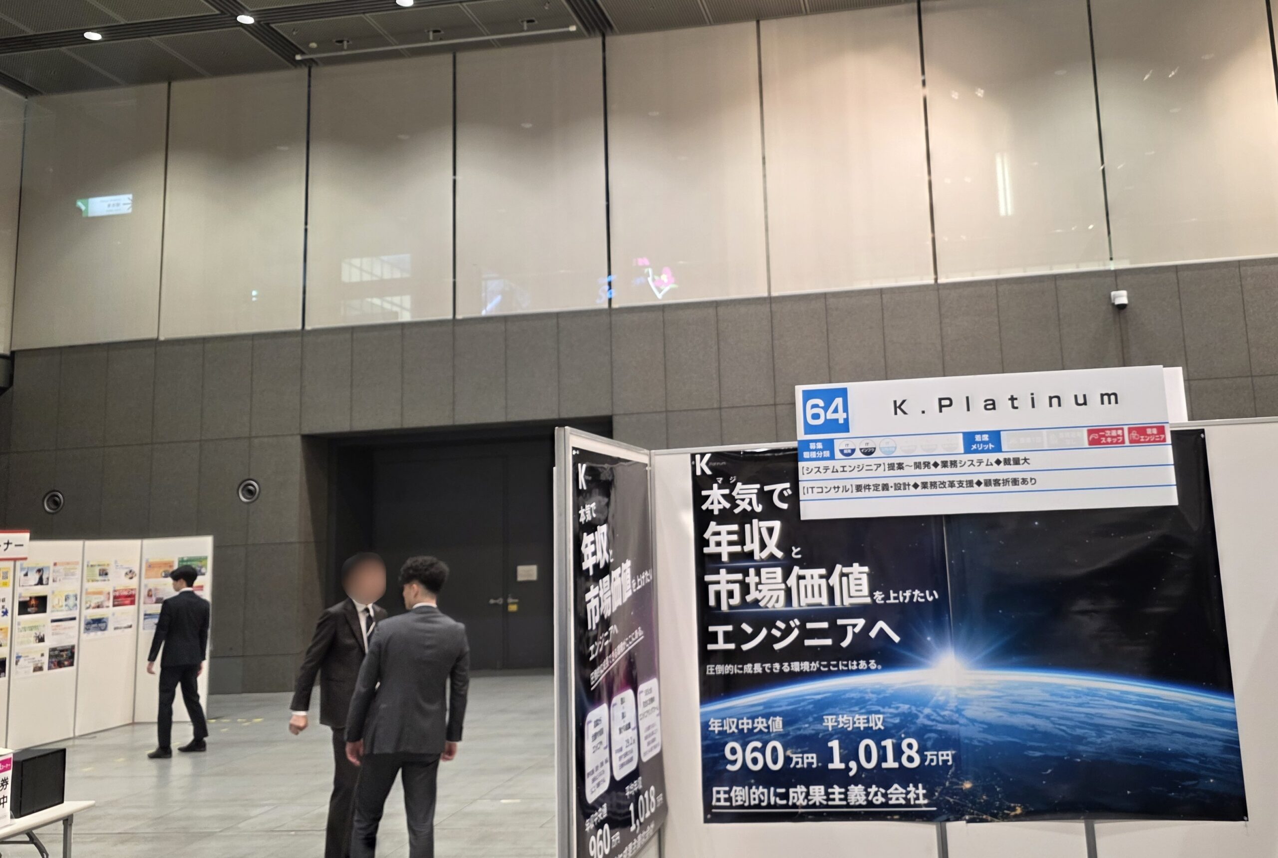近代的な屋内会場に、日本語のポスターが貼られた「K.Platinum」と書かれたリクルートブース。スーツ姿の男性2人がブースの近くで話している。.